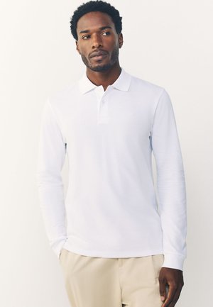 Mann mit kurzen lockigen Haaren, der ein weißes langärmeliges Poloshirt und beige Hosen trägt, steht mit einer Hand in der Tasche.