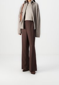 Maglione a coste beige sotto una giacca oversize grigia con zip, abbinati a pantaloni a campana marroni a vita alta e stivali con tacco marroni.