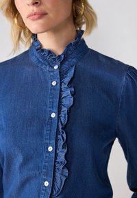 Blå denimskjorte med volangkrage og placket. Har hvite knapper og puffede ermer, og viser et jevnt, teksturert stoff.