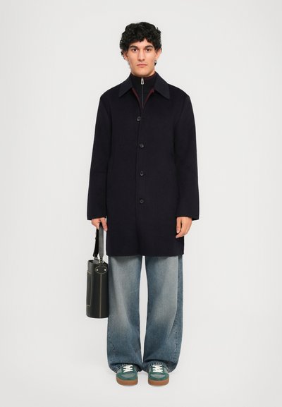 Man in black buttoned coat, loose faded blue jeans, green and white sneakers, holding black cylindrical bag against plain background.