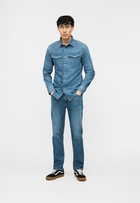 Νέος άνδρας στέκεται με τα χέρια στις τσέπες, φορώντας πουκάμισο από τζιν, τζιν παντελόνι και μαύρα αθλητικά παπούτσια με λευκές κορδόνια, σε απλό λευκό φόντο.