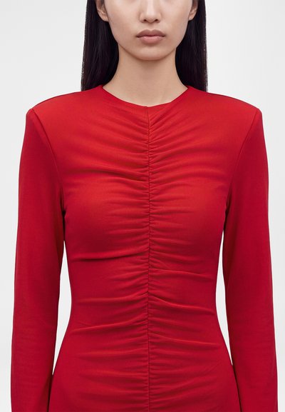 Femme portant un haut rouge ajusté à manches longues avec un détail froncé au centre devant.