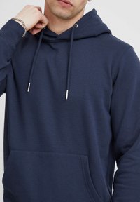 Sudadera de color azul marino hecha de tejido suave, con un bolsillo canguro, capucha con cordón y puntas metálicas, y puños acanalados.