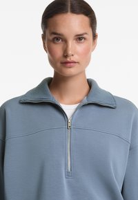 Modrá mikina na zips s rebrovaným golierom, vyrobená z hladkého materiálu a s polovičným zipsom vpredu. Má horizontálny ševový detail cez hrudník.