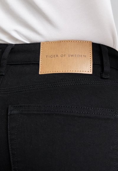 Schwarze Denim-Jeans mit einem braunen Lederlabel am Bund, auf dem der eingeprägte Text "TIGER OF SWEDEN" zu sehen ist. Minimalistisches Design mit sauberen Nähdetails.