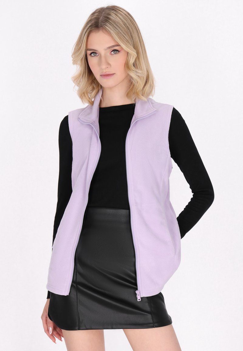 Gilet in pile viola chiaro con chiusura a zip e colletto alto, indossato sopra una maglietta nera a maniche lunghe, abbinato a una mini gonna in pelle nera.