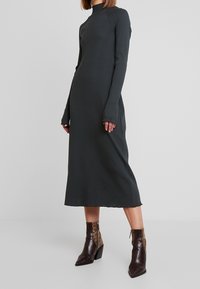 Robe côtelée gris foncé avec un col montant et des manches longues, arborant un ourlet effiloché. Associée à des bottines marron à motif.