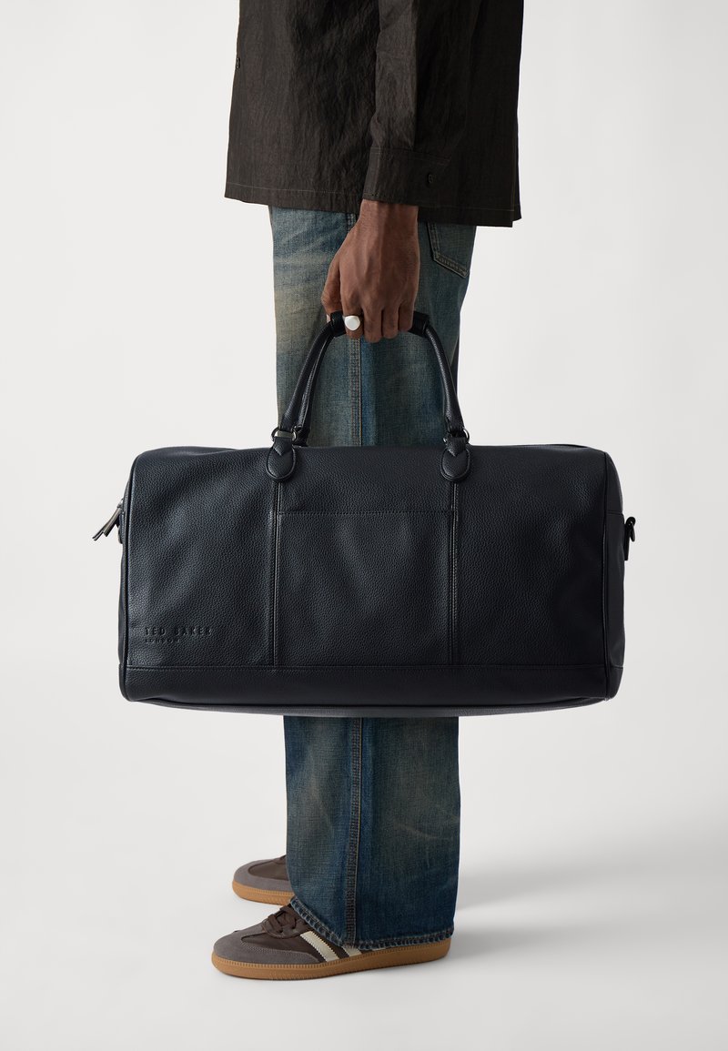 Black leather duffel bag with textured finish, dual top handles, and a front pocket. Features contrasting stitching and a zip closure.