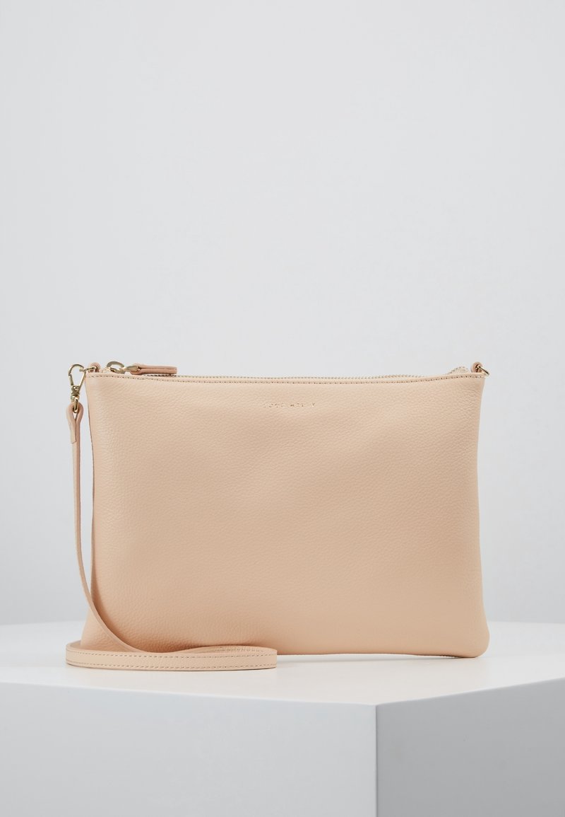 Pochette bracelet en cuir beige avec fermeture éclair dorée et sangle de poignet détachable, exposée sur un piédestal blanc sur fond gris clair.