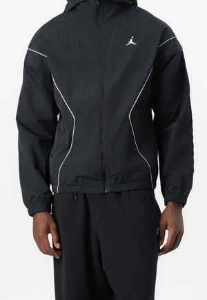 Person trägt eine schwarze Jacke mit Reißverschluss, weißen Paspeln und kleinem weißem Jumpman-Logo auf der linken Brust, kombiniert mit schwarzen Hosen.