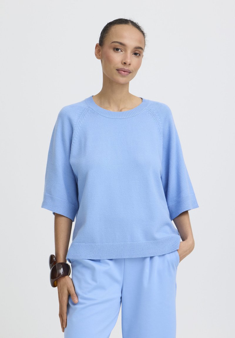 Femme portant un pull à manches courtes bleu clair et un pantalon assorti, debout avec une main dans la poche et un bracelet marron au poignet.