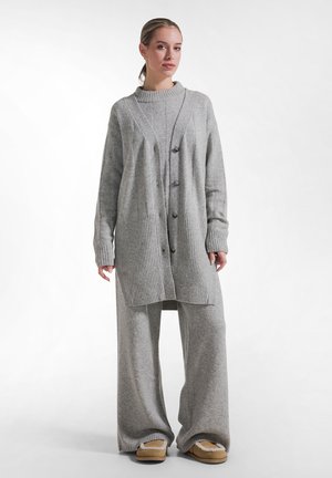 Kvinde iført løs grå strikket cardigan, matchende vidde bukser og lysebrune slip-on sko med hvid kant, stående mod hvid baggrund.
