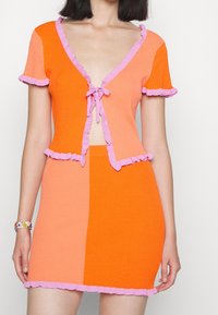Pletený dvoudílný outfit v oranžové a růžové barvě. Vrchní díl má výstřih do V a zavazování; oba kusy mají volánové okraje.