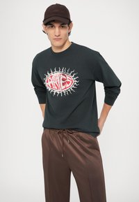 Cămașă neagră cu mâneci lungi și design grafic roșu, asortată cu pantaloni maro cu șnur. Materialul pare moale și cu o croială lejeră.