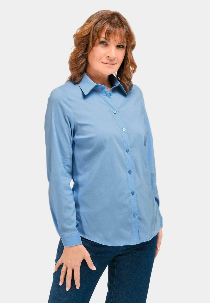 Hellblaues Hemd mit langen Ärmeln, Kragen und glatter Textur, kombiniert mit dunklen Jeans. Hat eine taillierte Passform und Knöpfe vorne.