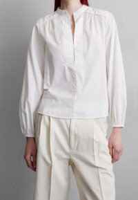 Valkoinen pitkähihainen pusero mandariinikauluksella, jossa on pintuck-yksityiskohtia ja suora helma. Yhdistetty vaaleanbeigeihin pleatattuihin housuihin.
