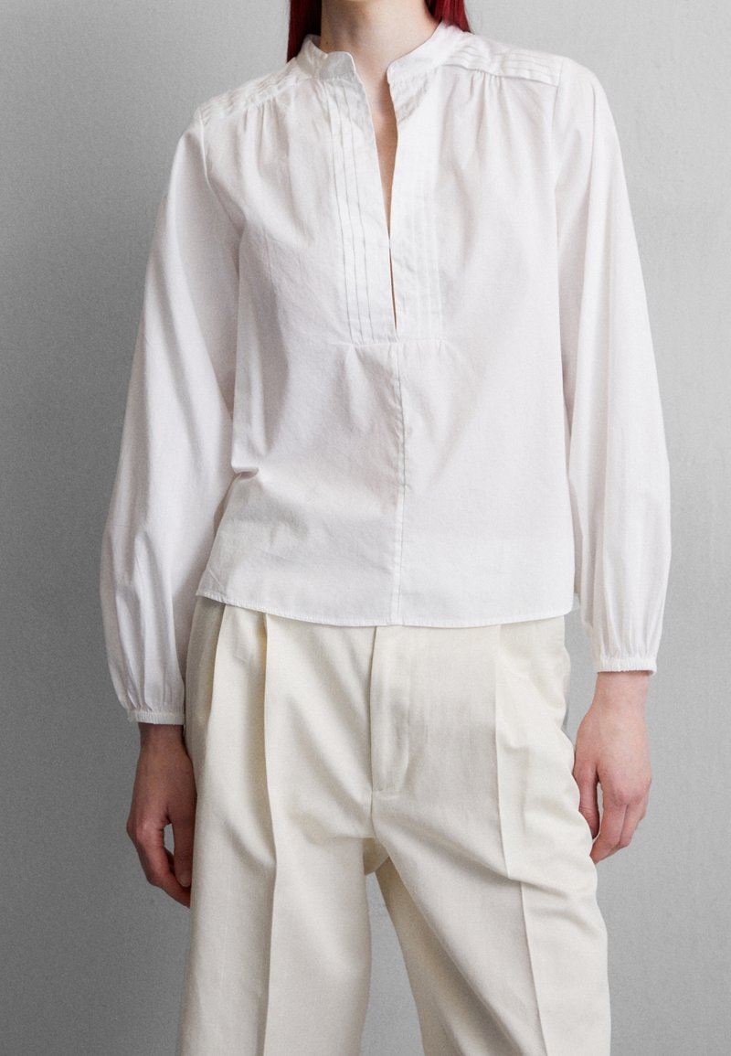 Valkoinen pitkähihainen pusero mandariinikauluksella, jossa on pintuck-yksityiskohtia ja suora helma. Yhdistetty vaaleanbeigeihin pleatattuihin housuihin.