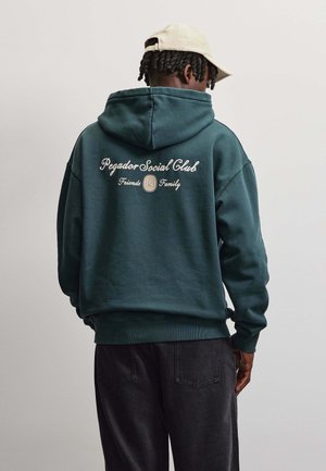 Persona che indossa una felpa con cappuccio verde scuro con il testo "Pegador Social Club Friends Family" e un cappellino beige, rivolta di spalle su uno sfondo semplice.