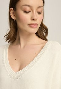 Biała sweter z dzianiny z dekoltem w kształcie litery V, z prążkowanymi wykończeniami i subtelną fakturą. Materiał jest miękki o delikatnym, lekkim wyglądzie.