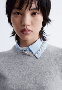 Grauer Strickpullover über einem hellblauen Hemd mit Kragen, ausgestattet mit einer silbernen Kettenhalskette. Die Textur wirkt weich und glatt.