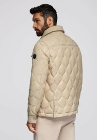 Uomo con capelli scuri e barba che indossa una giacca trapuntata beige e pantaloni chiari, rivolto di spalle e guardando di lato.