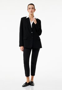 Sort blazer med krave, to frontlommer og enkeltknaplukning, parret med skræddersyede sorte bukser og sorte loafers.