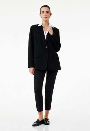 Blazer negro con cuello muesca, dos bolsillos frontales y cierre de un solo botón, combinado con pantalones negros ajustados y mocasines negros.