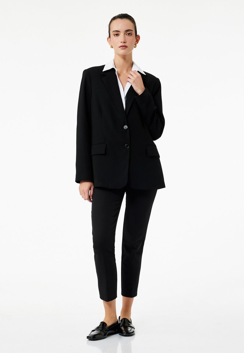 Sort blazer med krave, to frontlommer og enkeltknaplukning, parret med skræddersyede sorte bukser og sorte loafers.