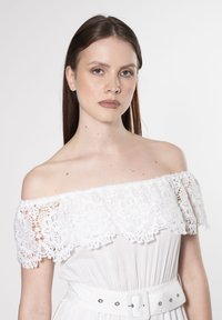 Weißes schulterfreies Kleid mit einem Spitzenrüschendetail am Ausschnitt, tailliert mit einem weißen Gürtel und auffälligen Lochmuster-Akzenten.