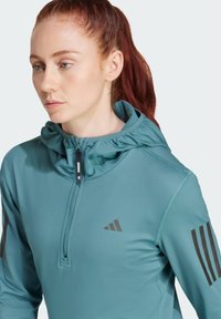 Tealfärgad träningshoodie med dragkedja vid halsen, samlad huva och svarta tre-stripes detaljer på ärmarna. Slätt, lättviktsmaterial.