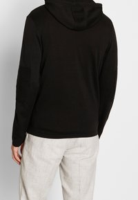 Uomo che indossa una felpa con cappuccio nera con toppe a coste sui gomiti e pantaloni beige chiaro dal tessuto strutturato, mostrato di spalle su uno sfondo semplice.