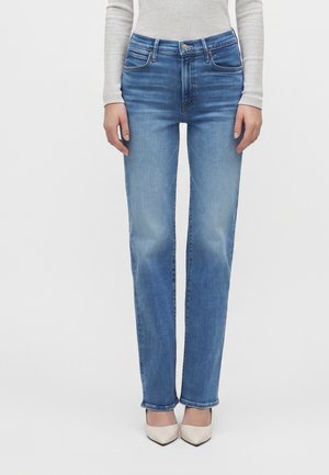 Vrouw die lichtblauwe jeans met rechte pijpen, een beige top met lange mouwen en witte pumps met puntige neus draagt en staat tegen een effen achtergrond.