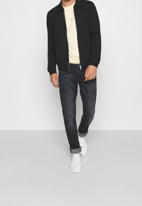 Musta vetoketjullinen takki vaaleankeltaisen paidan päällä, yhdistetty tummiin farkkuihin ja valkoisiin tennareihin. Rentoon istuvuuteen sopiva yksinkertainen muotoilu ilman näkyviä kuvioita.