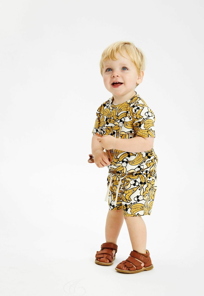 Kortärmad outfit med ett bananmönster i gult och svart, åtföljd av bruna sandaler, tillverkad av lättviktsmaterial.