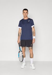Tumesinine sportlik T-särk valgete varrukate ja mustade lühikesed püksid, millel on oranž logo, valged sokid ja valged tossud, hoides tennisereketit.