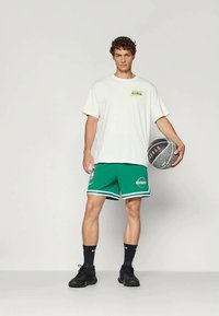 Ung mann som holder en basketball, iført hvit Nike t-skjorte, grønne Boston Celtics shorts, svarte Nike sokker og svarte joggesko.