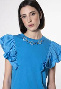 Top blu brillante con maniche a volant e collana decorativa a catena con anelli geometrici; tessuto morbido e design aderente.