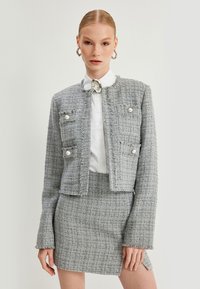 Harmaa tweed-takki, jossa on rispaantuneet reunat, neljä etutaskua ja helmiakcentit. Yhdistetty vastaavaan istuvaan hameeseen. Valkoinen kauluspaita alla.