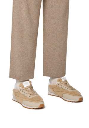 Beige Sneakers mit einem strukturierten, flauschigen Obermaterial, hellbeige Wildlederelementen und einer weißen Sohle. Kombiniert mit gerippten beigen Hosen und hellen Socken.