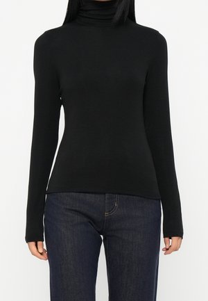 Žena v černém přiléhavém roláku s dlouhým rukávem a tmavě modrými džíny stojící před jednobarevným pozadím.