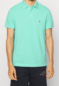 Mintgrön pikétröja i bomull, korta ärmar, två knappslå, logotyp på vänster bröst. Bärs med marinblå sportshorts.
