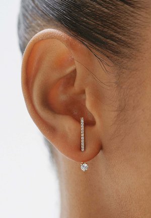 Primer plano de una oreja con un pendiente de barra vertical de oro con pequeños diamantes y un pendiente de diamante redondo colgando debajo.