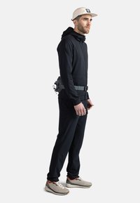 Schwarze Sportjacke mit einem Frontreißverschluss, kombiniert mit schwarzen Hosen und grauen Turnschuhen. Ein grauer Gürtel mit angebrachten Utensilien wird um die Taille getragen.