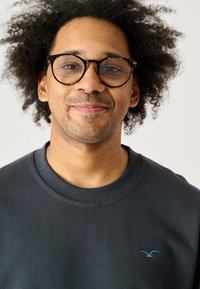 Grauer Pullover mit Rundhalsausschnitt und gerippter Textur, mit einem kleinen türkisfarbenen Vogel-Logo auf der linken Brust. Weißer Hintergrund.