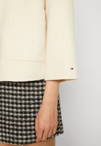 Beige wollen jas met een zak en logo-detail, gecombineerd met een zwart-witte geruite rok, die textuur en contrasterende patronen toont.