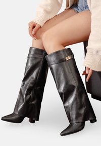 Bottes noires montantes en cuir brillant, dotées d'un rebord replié, d'une fermeture en accent doré, d'un bout pointu et d'un talon épais bas.