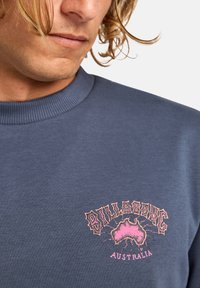 Blue sweatshirt with a ribbed neckline featuring a pink graphic design of Australia and the text "BILLABONG AUSTRALIA" in bold font.