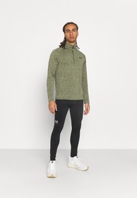 Grønn, langermet sportsgenser med glidelås i kragen, sammen med svarte kompresjonsleggings. Hvite joggesko med teksturert såle og logo.