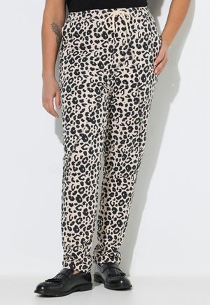 Persona con pantalones beige de estampado de leopardo y cintura con cordón, zapatos mocasines negros, mano apoyada en la cadera, sobre fondo blanco.