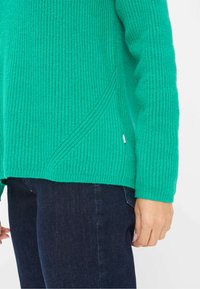 Grøn strikket sweater med et tekstureret mønster, der har en skrå sømdetalje og ribbede manchetter, parret med mørkeblå jeans.